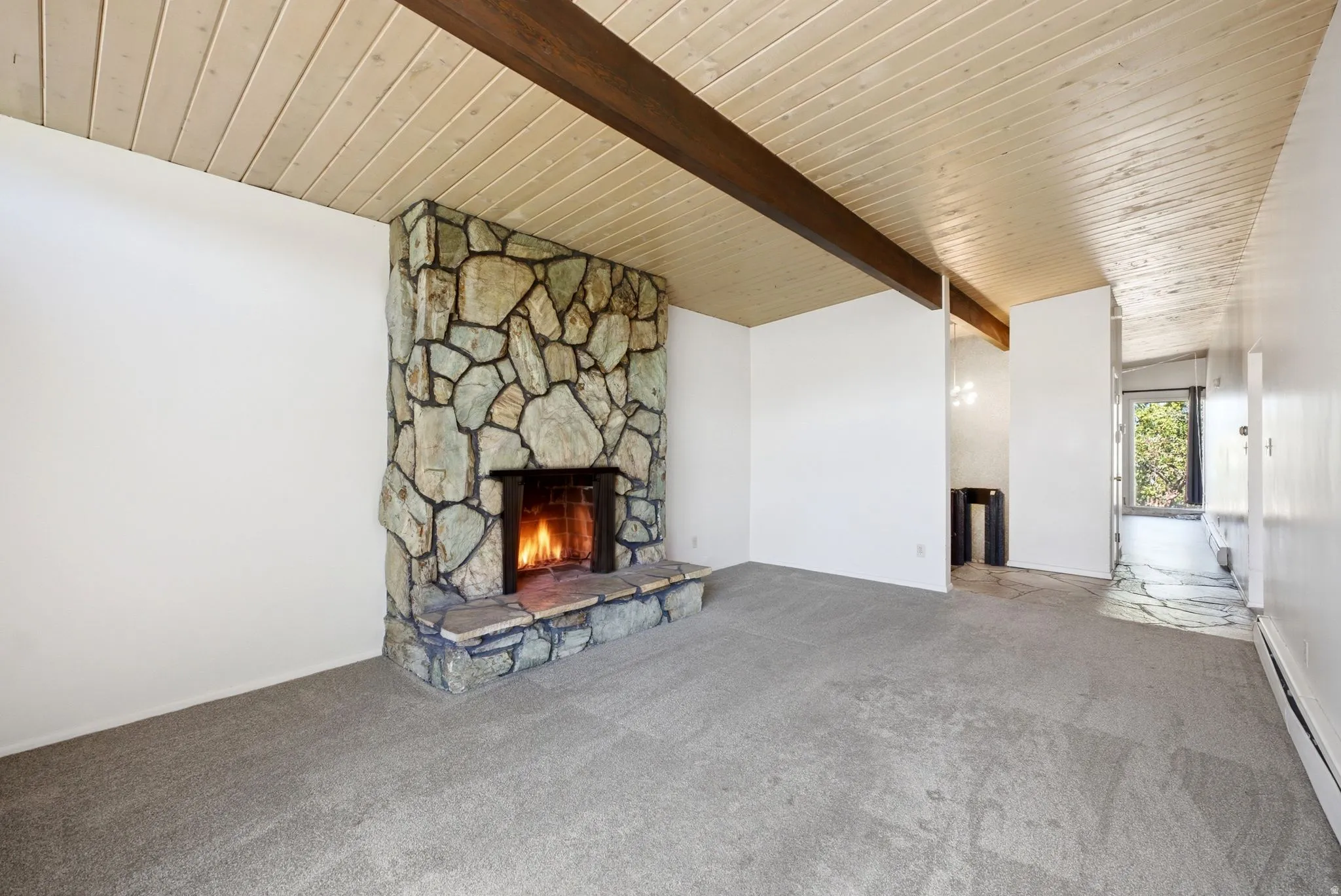 Stone fireplace with hearth, exposed wood beam, wood-panel vaulted ceiling, and grey carpet flooring