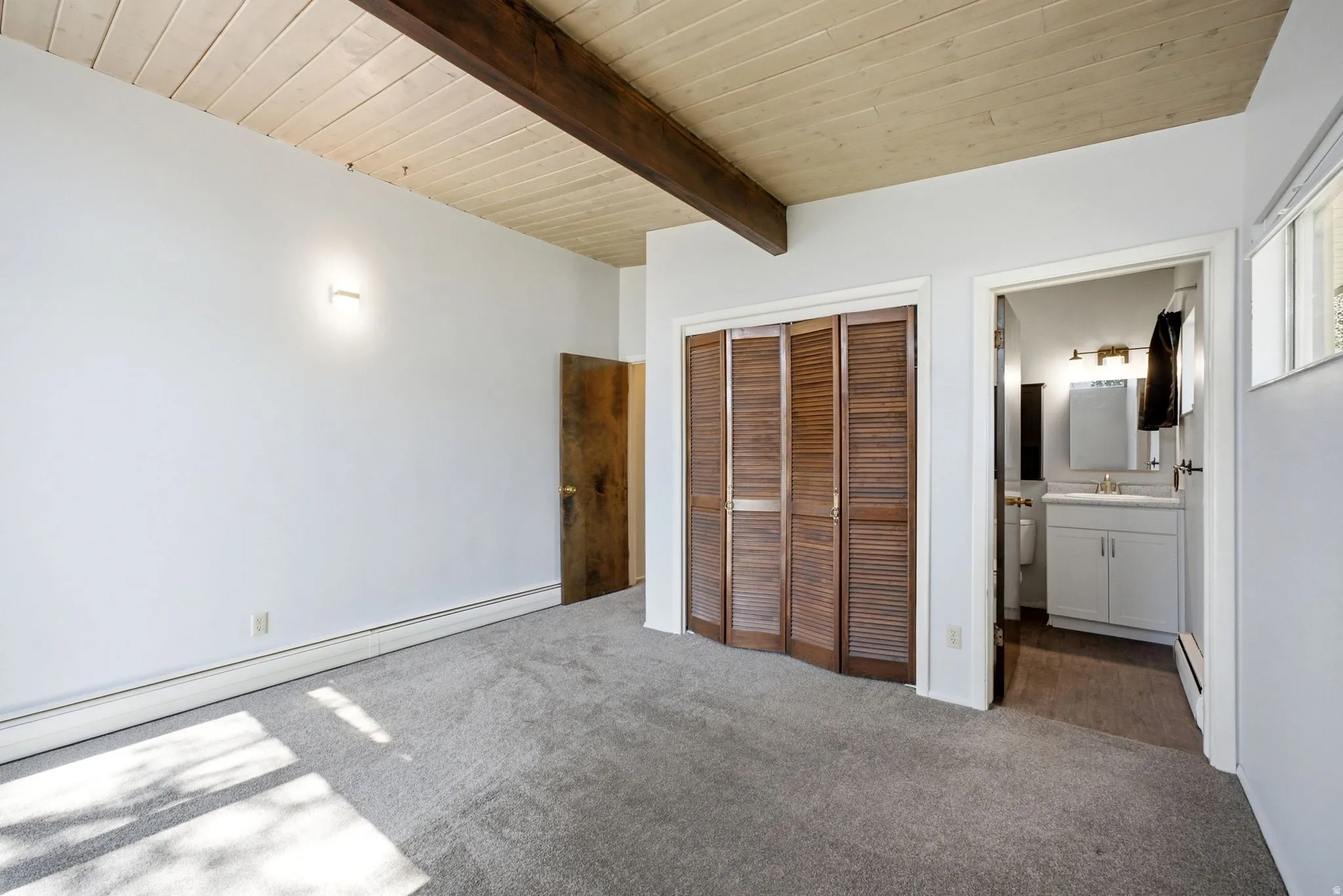 Exposed ceiling beams, wood panel ceiling, wall-mounted light fixture, louvered closet doors, and an attached bathroom with a vanity