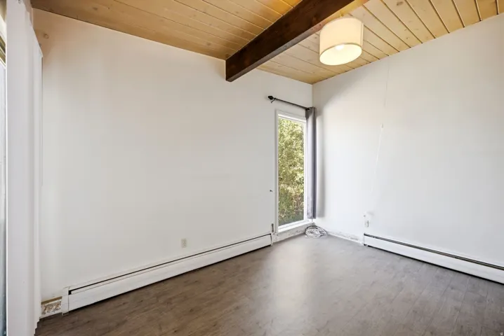 Wood-finish flooring with baseboard heating, vertical window, and wood panel ceiling with exposed beam and flush-mount light fixture