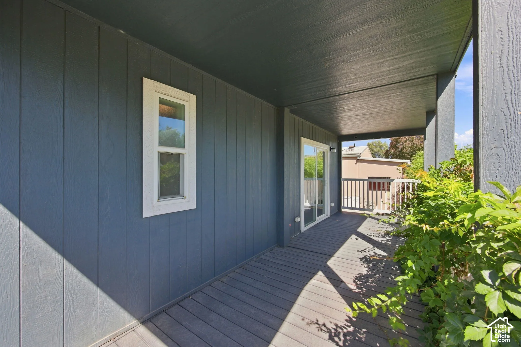 Covered outdoor deck with a sliding glass door, white window trim, and vertical panel siding