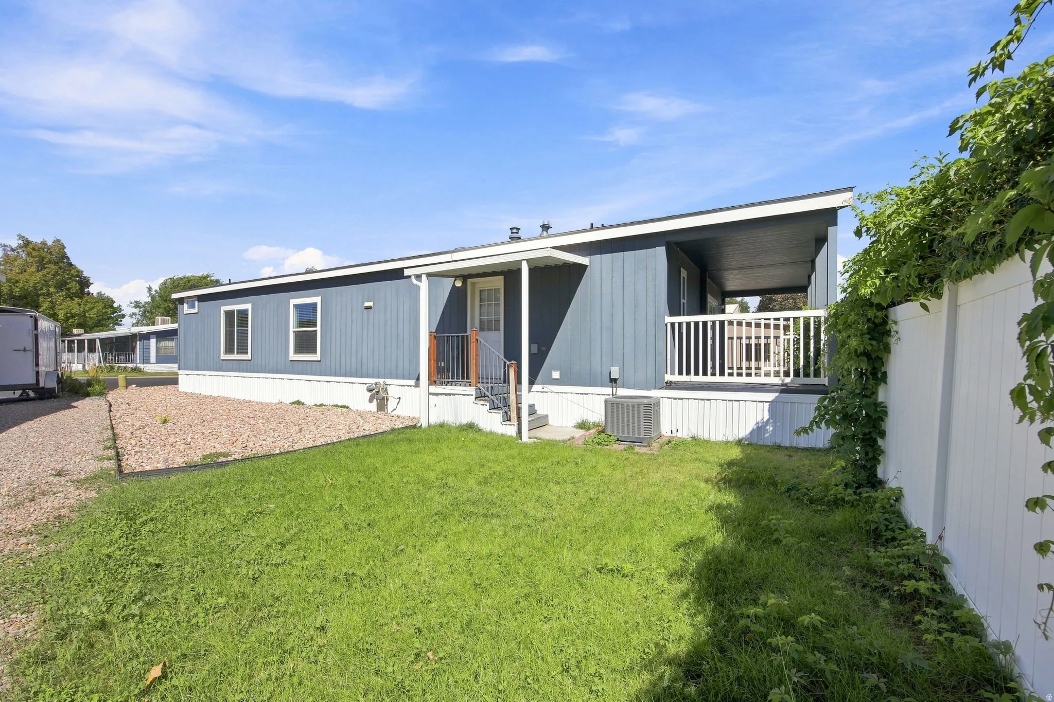 Modern manufactured home featuring a covered front porch with white railings, metal siding exterior in gray and white tones, large windows for natural light, and a spacious manicured lawn with mature landscaping and privacy fencing along the perimeter