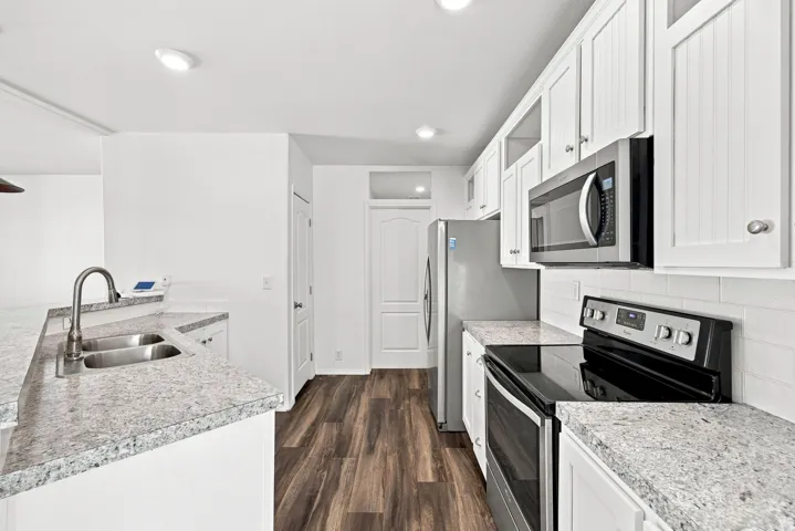 Modern kitchen featuring white cabinetry with glass-front uppers, stainless steel appliances including a double wall oven and microwave, granite countertops, an undermount sink with contemporary faucet, and wood-finish flooring throughout the galley layou