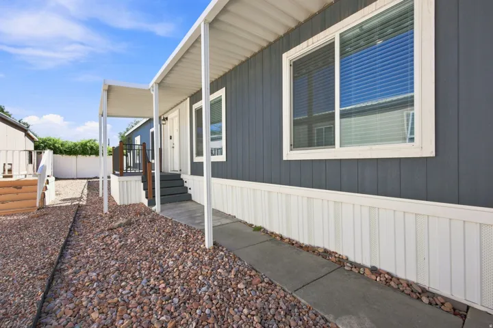 Modern manufactured home featuring a sleek two-tone exterior with dark gray and white metal siding, expansive front-facing window with horizontal blinds, covered entry porch with white trim and railing, and a gravel landscaped yard with concrete walkway a