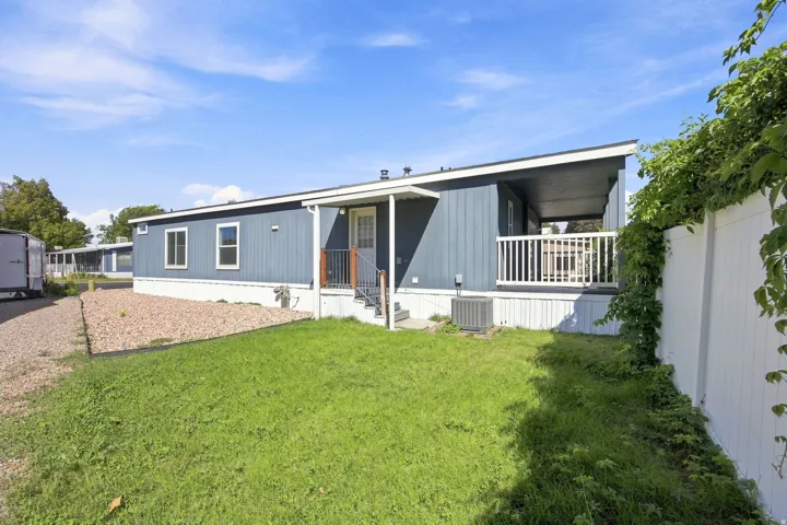 Modern manufactured home featuring a covered front porch with white railings, metal siding exterior in gray and white tones, large windows for natural light, and a spacious manicured lawn with mature landscaping and privacy fencing along the perimeter