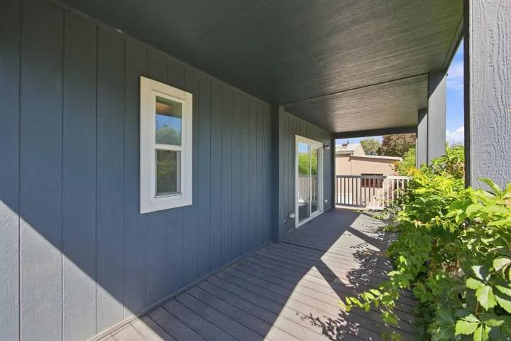 Covered outdoor deck with a sliding glass door, white window trim, and vertical panel siding
