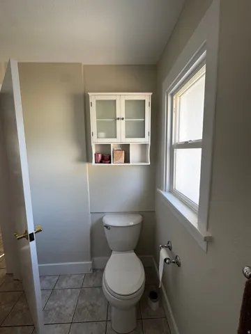 Two-piece toilet with a wall-mounted storage cabinet, window with white trim, and tiled flooring