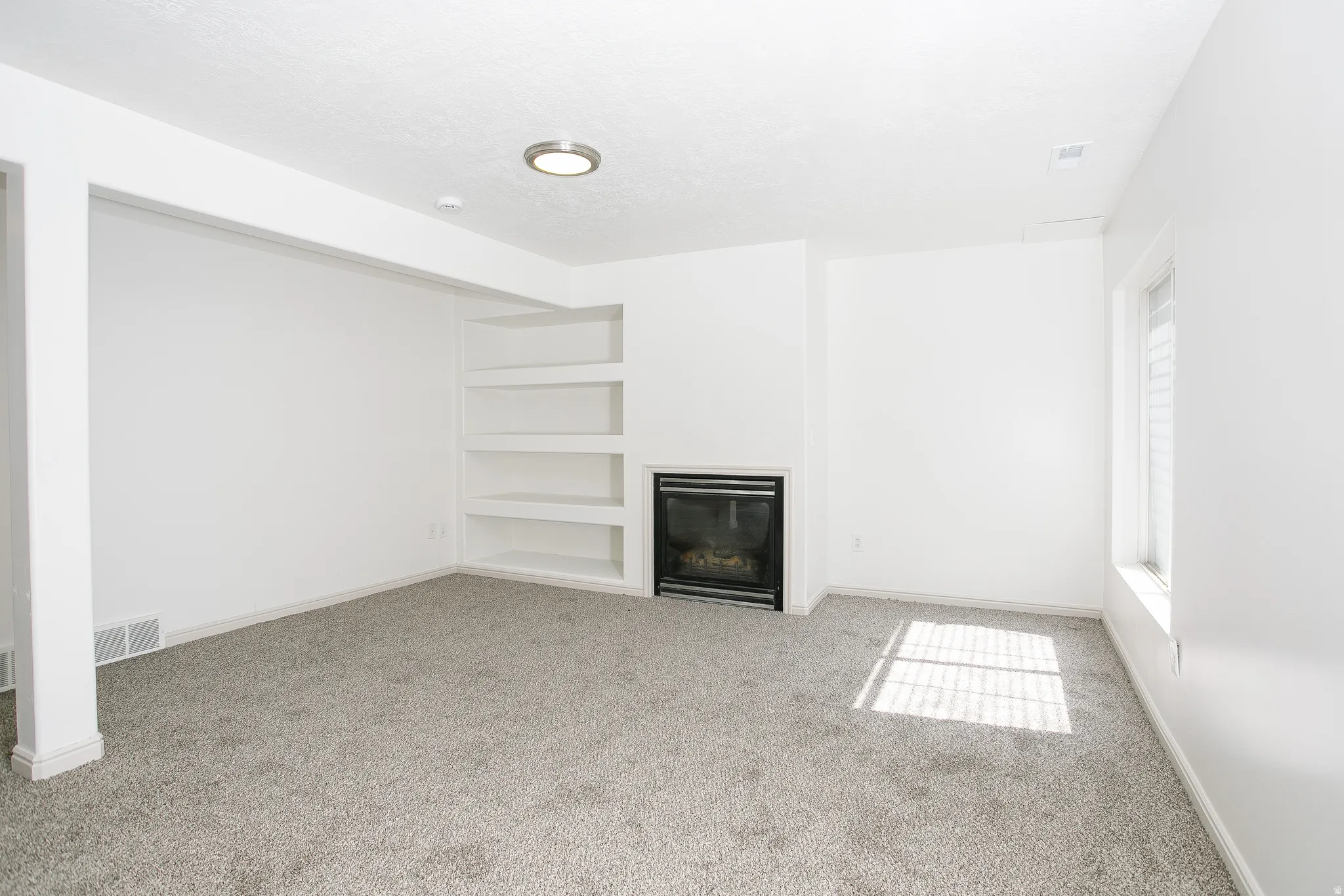Recessed fireplace with black surround, built-in shelving, gray carpeting, and a window with blinds