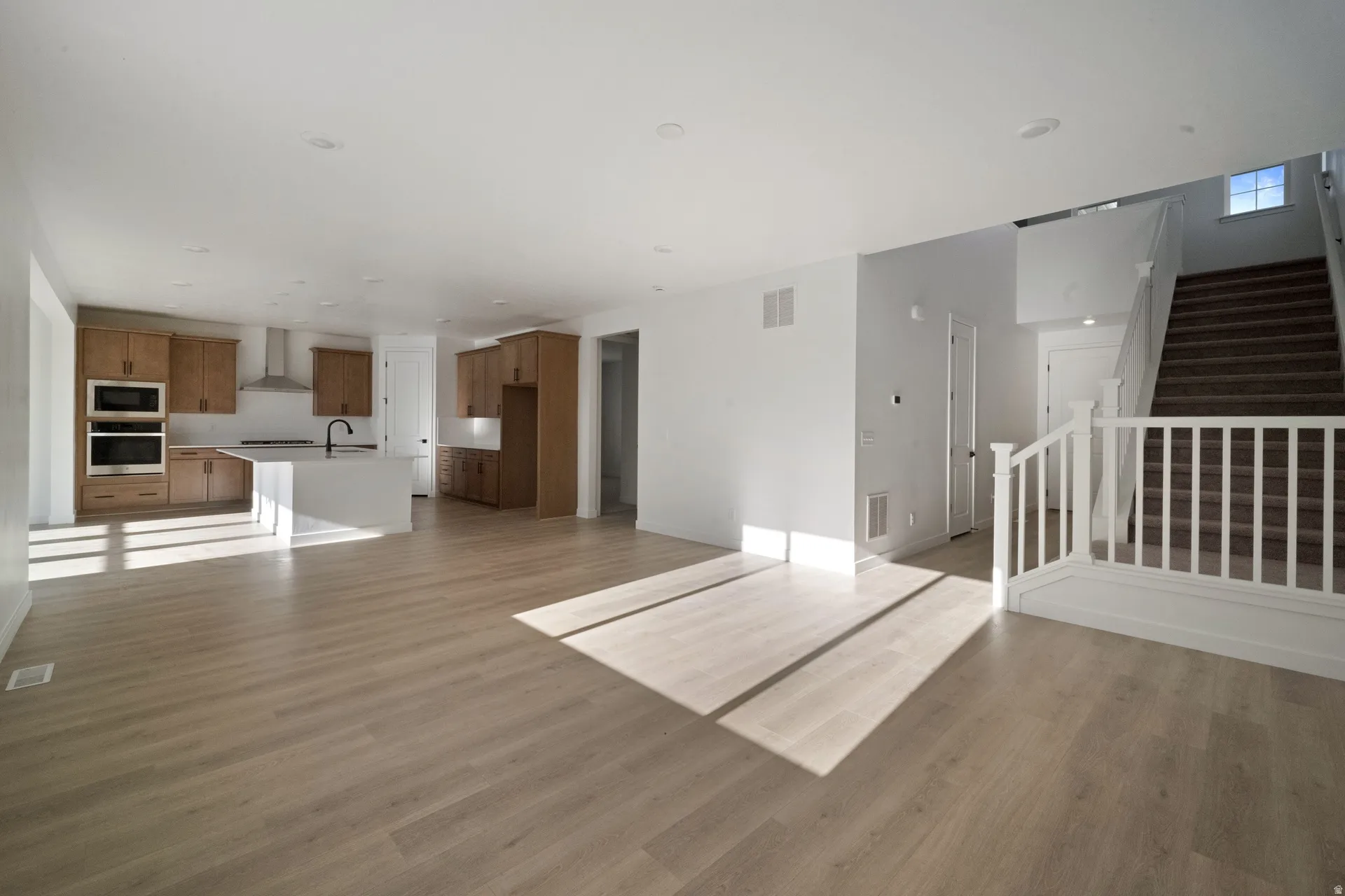 Open-concept living space featuring wood-finish flooring, recessed lighting, a kitchen island, double ovens, and a staircase with white balusters