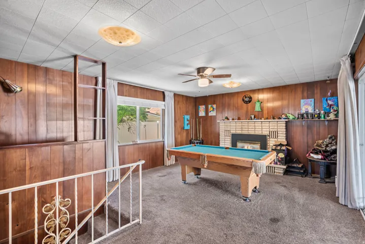 Wood paneling throughout, brick fireplace, built-in shelving, white railing, and ceiling fan