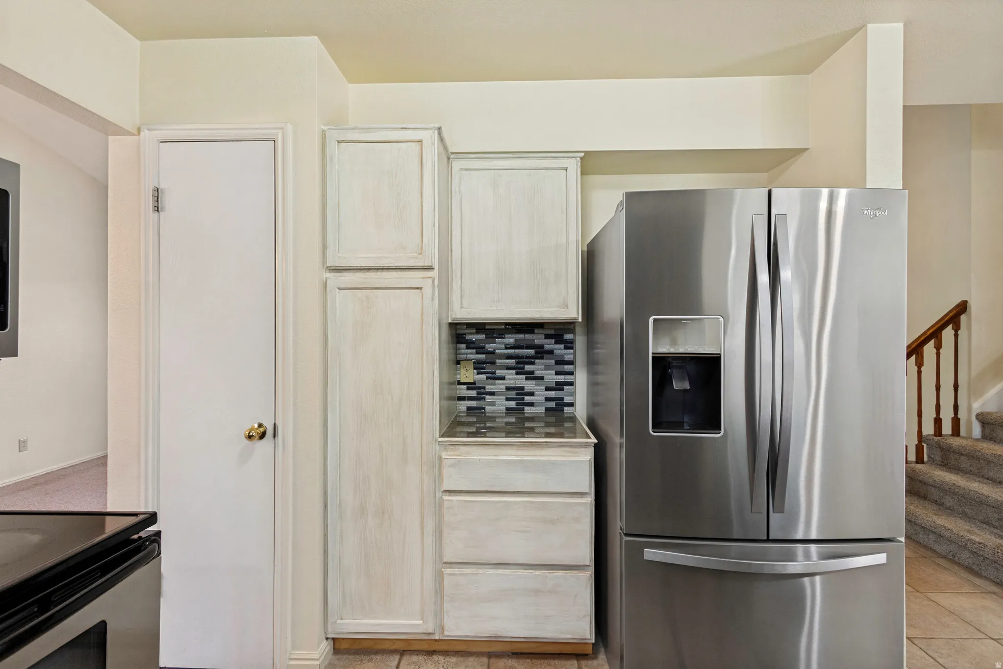 Stainless steel French door refrigerator, mosaic tile backsplash, light-toned cabinetry, and carpeted stairs with wood railing