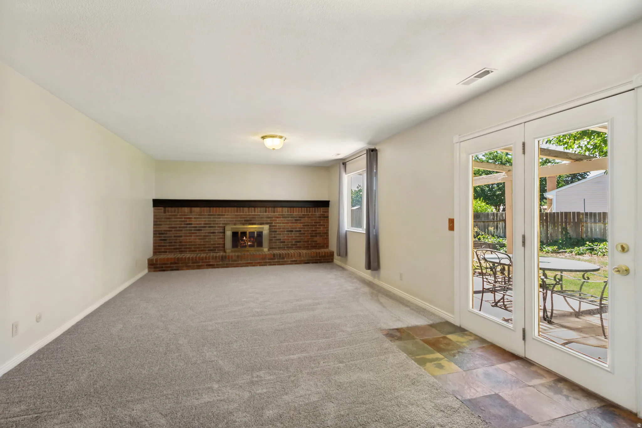 Brick fireplace with a brass firebox, wall-to-wall carpeting, window with drapery, double glass-paneled doors, and multi-color slate tile flooring