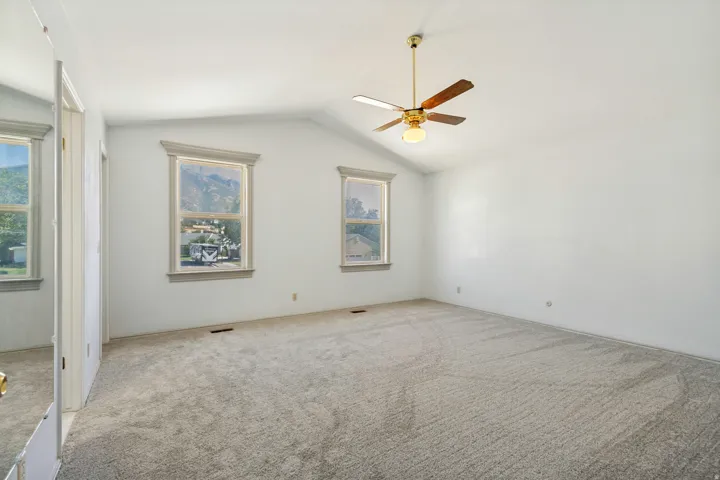 Primary Bedroom - Vaulted ceiling with a ceiling fan light fixture, two windows with decorative trim, and wall-to-wall carpeting