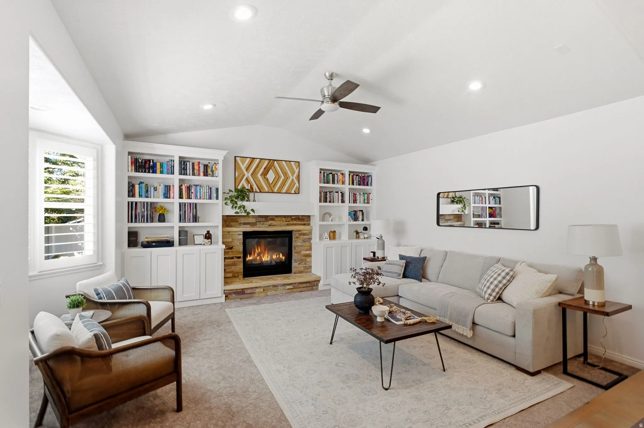 Vaulted ceiling with recessed lighting, stone-clad fireplace, integrated built-in shelving, window with plantation shutters, and ceiling fan
