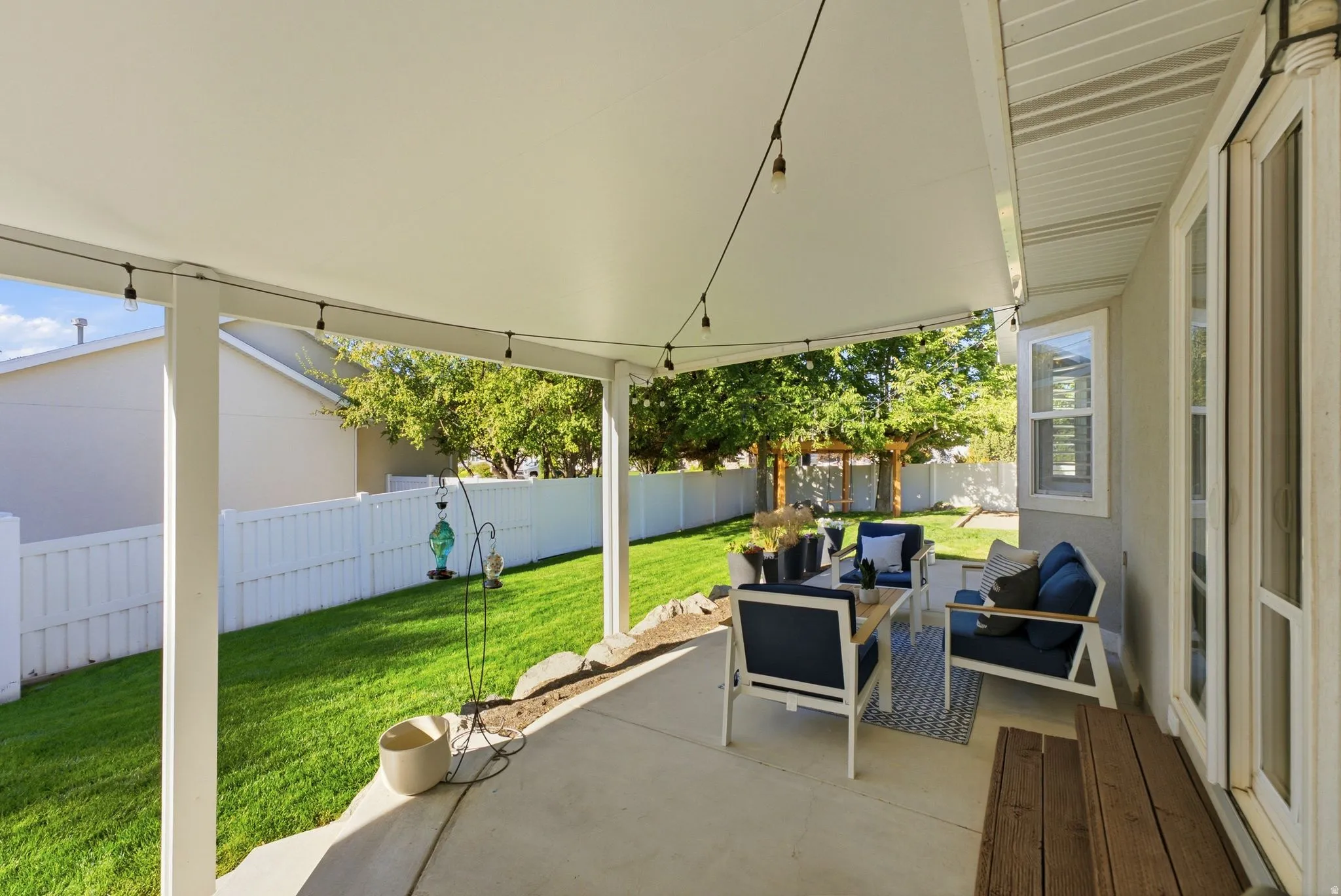 Covered concrete patio with string lighting, expansive lawn, and white privacy fencing
