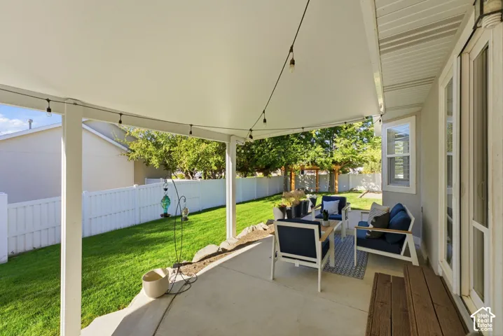 Covered concrete patio with string lighting, expansive lawn, and white privacy fencing