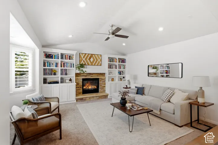 Vaulted ceiling with recessed lighting, stone-clad fireplace, integrated built-in shelving, window with plantation shutters, and ceiling fan