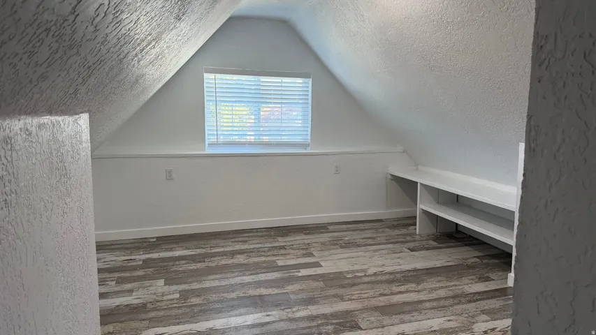 Attic space featuring wood-finish flooring, an angled ceiling, a centered window with blinds, and built-in shelving