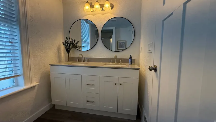 Dual vanity with integrated sinks, contemporary wall-mounted light fixture, two circular mirrors, and wood-finish flooring