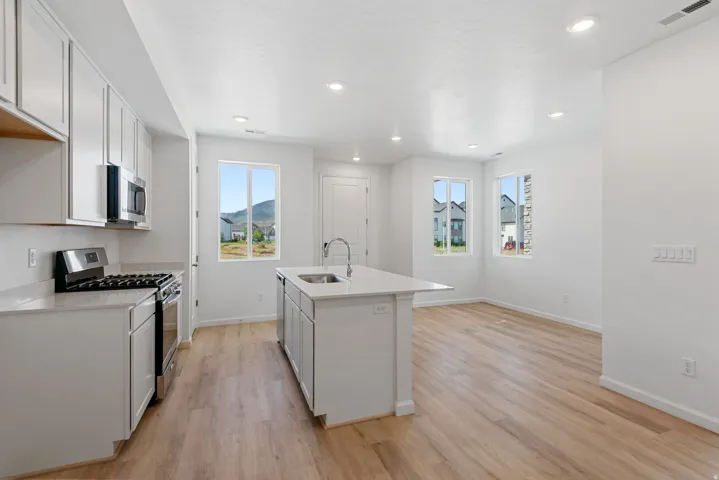 Recessed lighting illuminates the open concept kitchen featuring wood-finish flooring, white cabinetry, a central island with an undermount sink, and stainless steel appliances