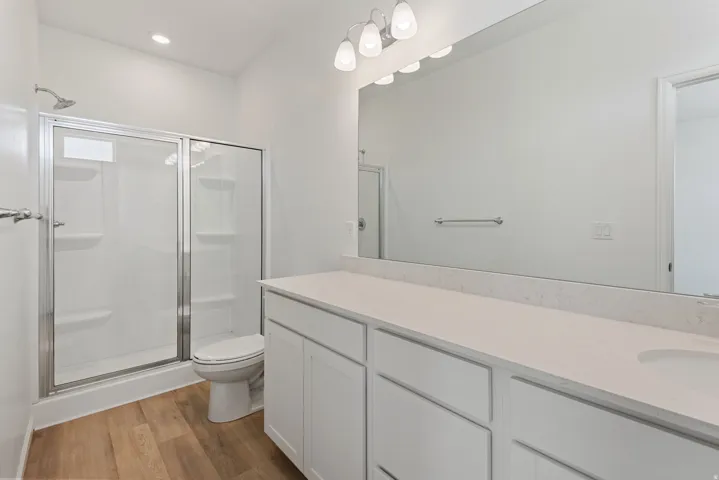 Spacious bathroom featuring a white dual vanity with undermount sinks, a large mirror, wood-finish flooring, and a glass-enclosed shower