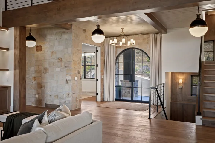 Exposed wood beams, stone accent wall, wood-finish flooring, arched black-framed glass door, and a staircase with a metal railing