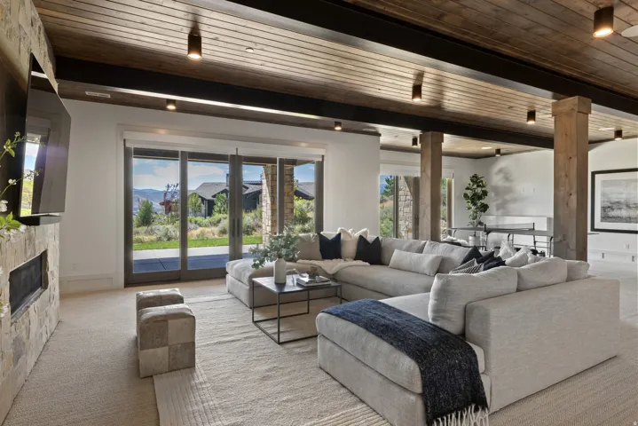 Spacious living area featuring a stacked stone fireplace, wood-plank ceiling, exposed wood beams, and a wall of glass doors opening to the exterior