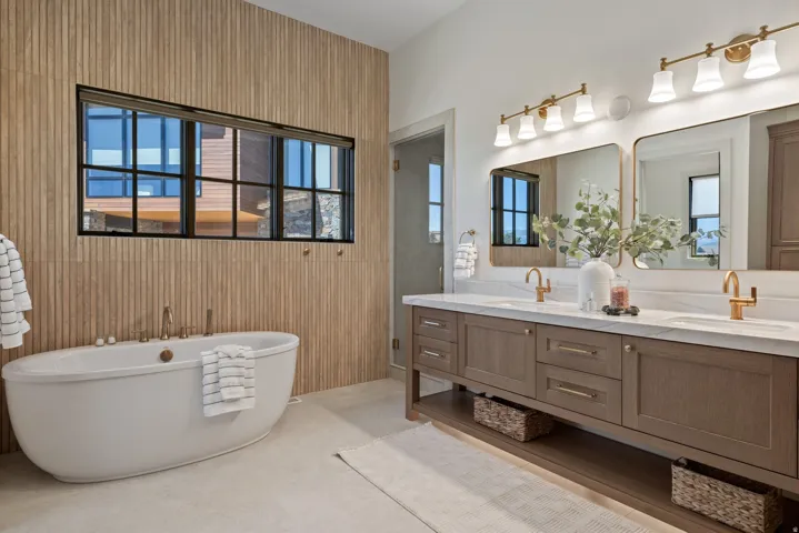 Freestanding soaking tub with wood-finish accent wall, dual vanity with undermount sinks, and gold-tone fixtures