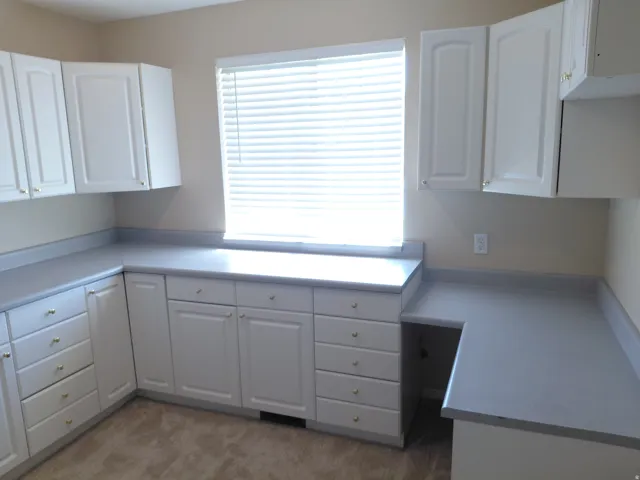 Extensive white cabinetry with brushed gold hardware, ample gray countertop space, built-in desk area, and a large window with blinds