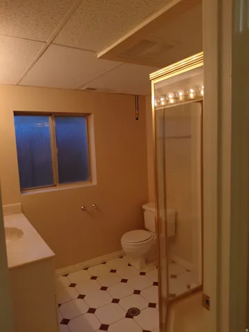 Bathroom with drop ceiling, window, vanity with integrated sink, toilet, and shower enclosure with gold-tone trim