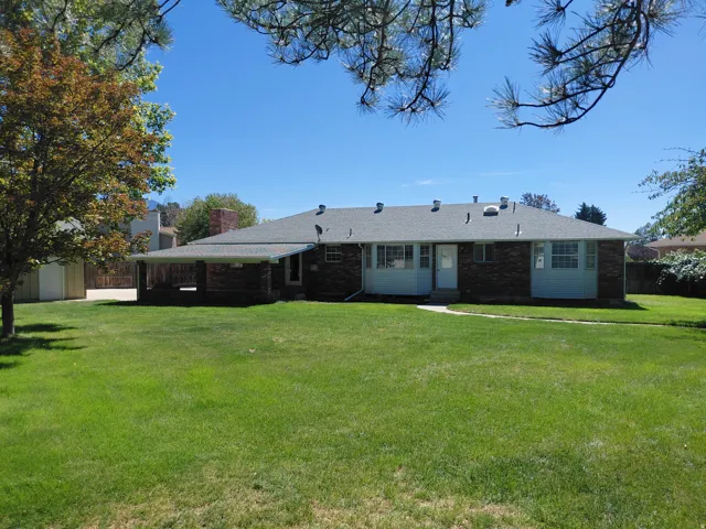 Expansive rear yard with a green lawn, mature trees, and a brick exterior home featuring a covered patio, a shed, and a paved drive