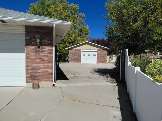 Brick-clad exterior with attached garage, concrete driveway, white vinyl fencing, and detached garage structure