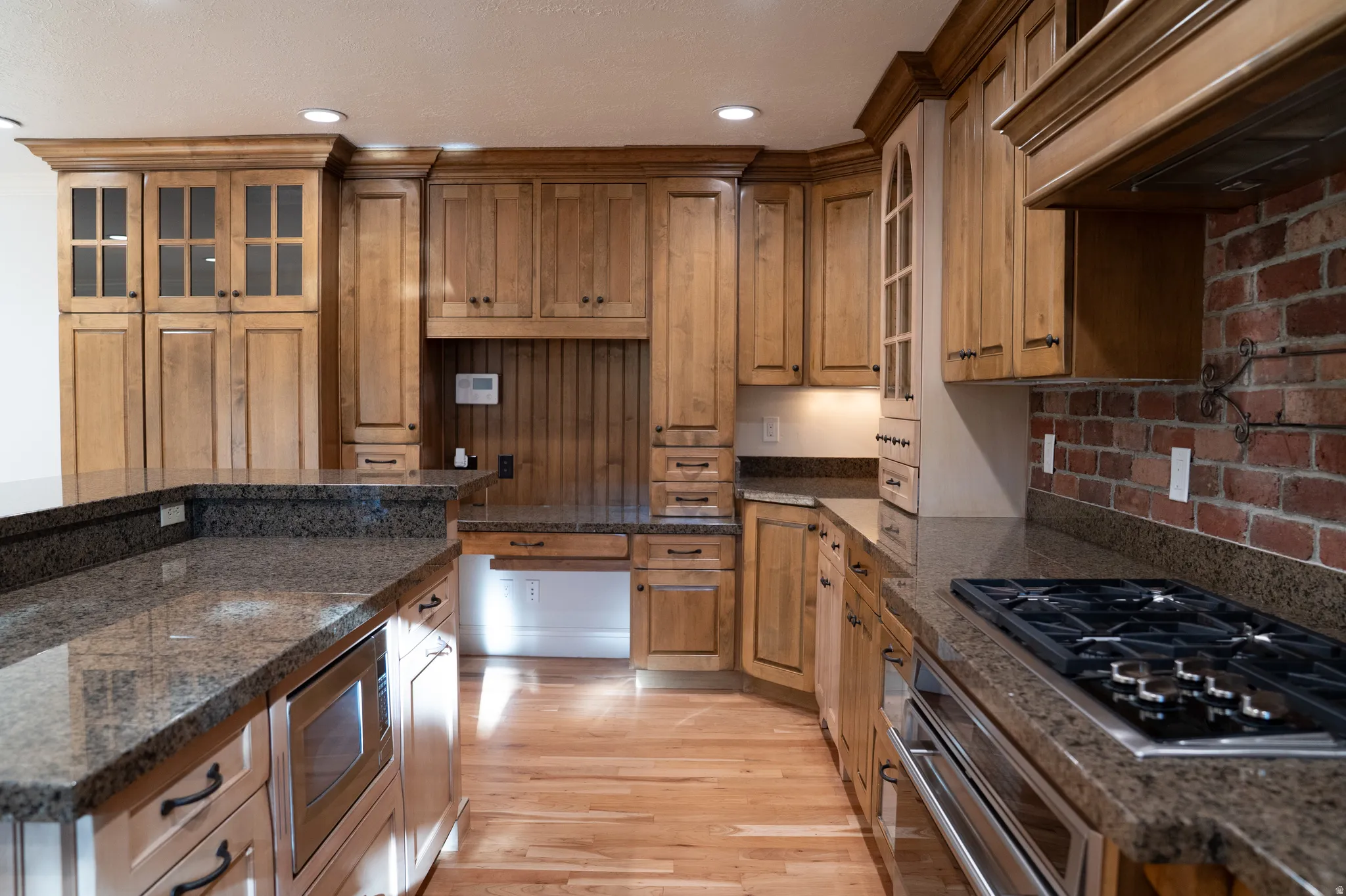 Wood-finish cabinetry with crown molding, dark granite countertops, hardwood flooring, brick-finish backsplash, and stainless steel appliances