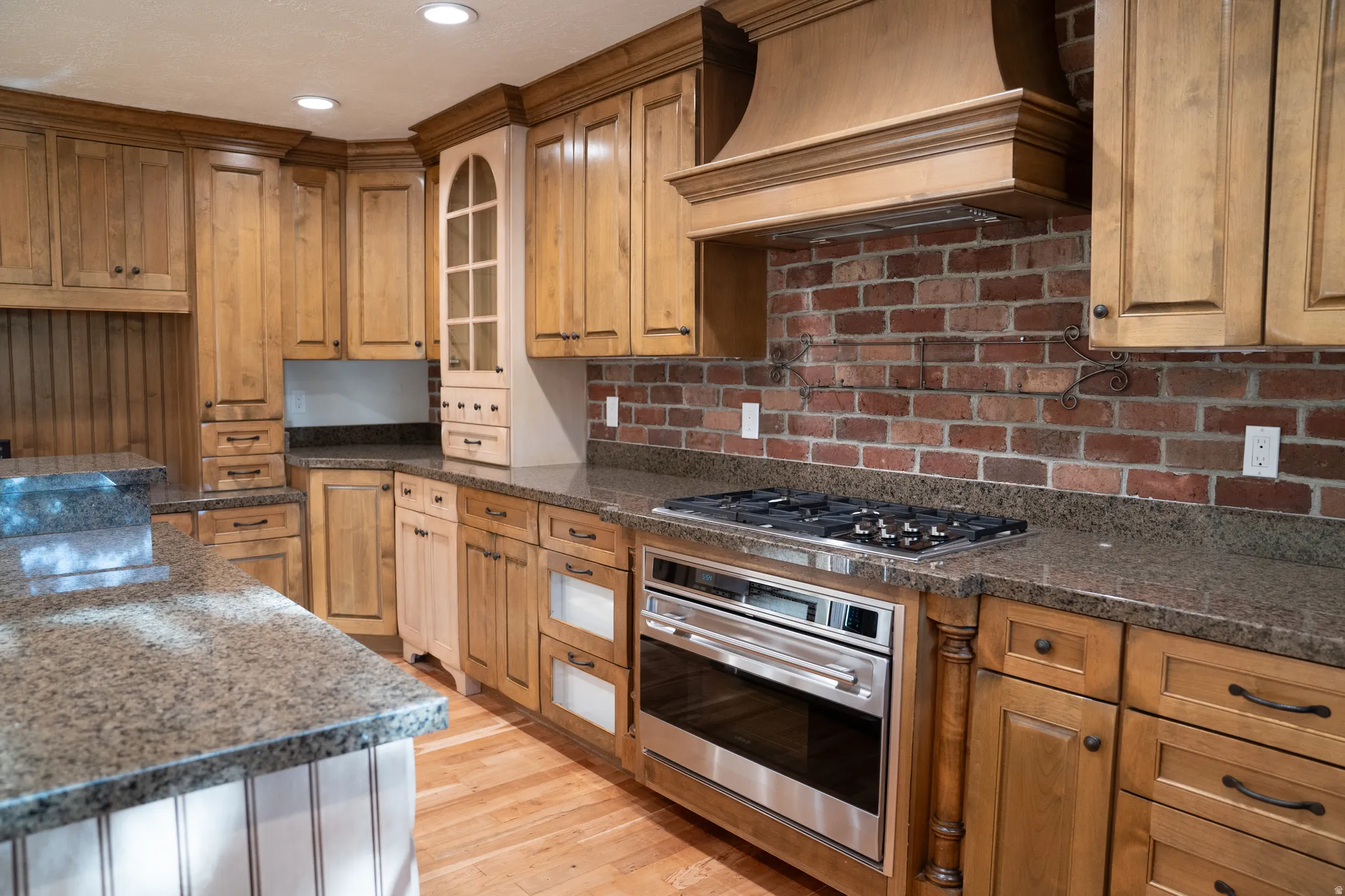 Wood-finish cabinetry with dark hardware, granite-style countertops, stainless steel appliances, brick-style backsplash, and wood-finish flooring