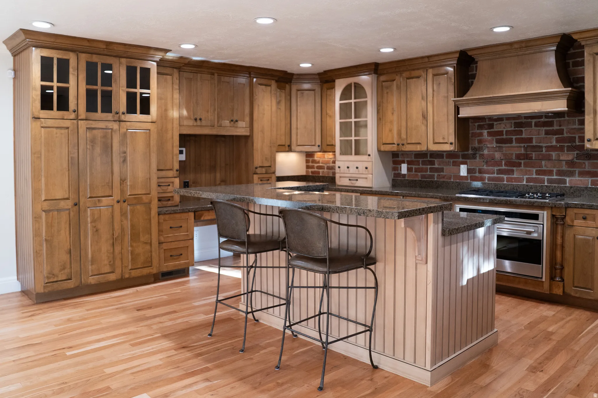 Wood-finish flooring, natural wood cabinetry, island with beadboard paneling, integrated stainless steel oven, and brick backsplash