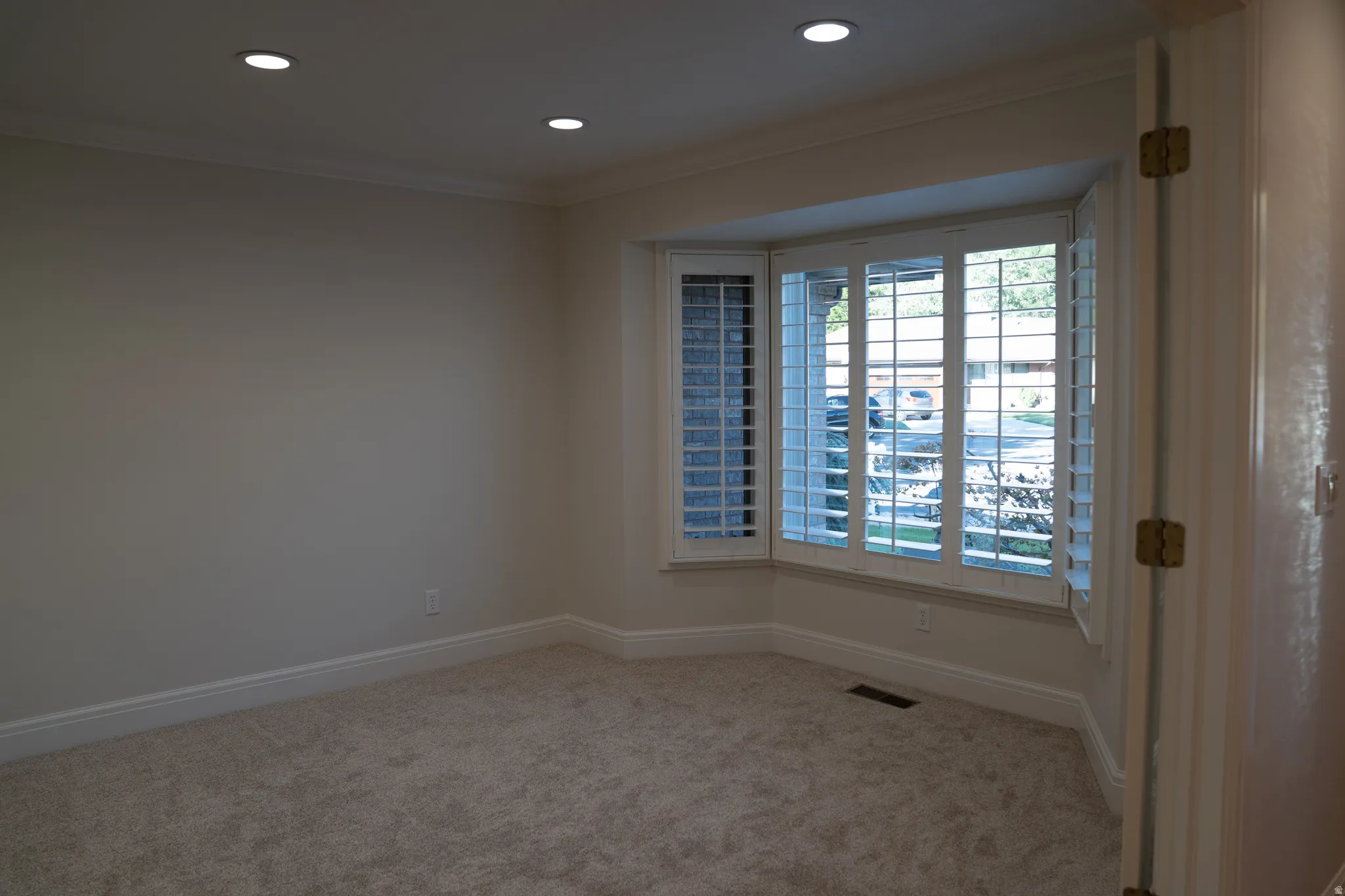Recessed lighting illuminating the room, crown molding accenting the ceiling, a bay window with plantation shutters, plush carpeting, and crisp white baseboards