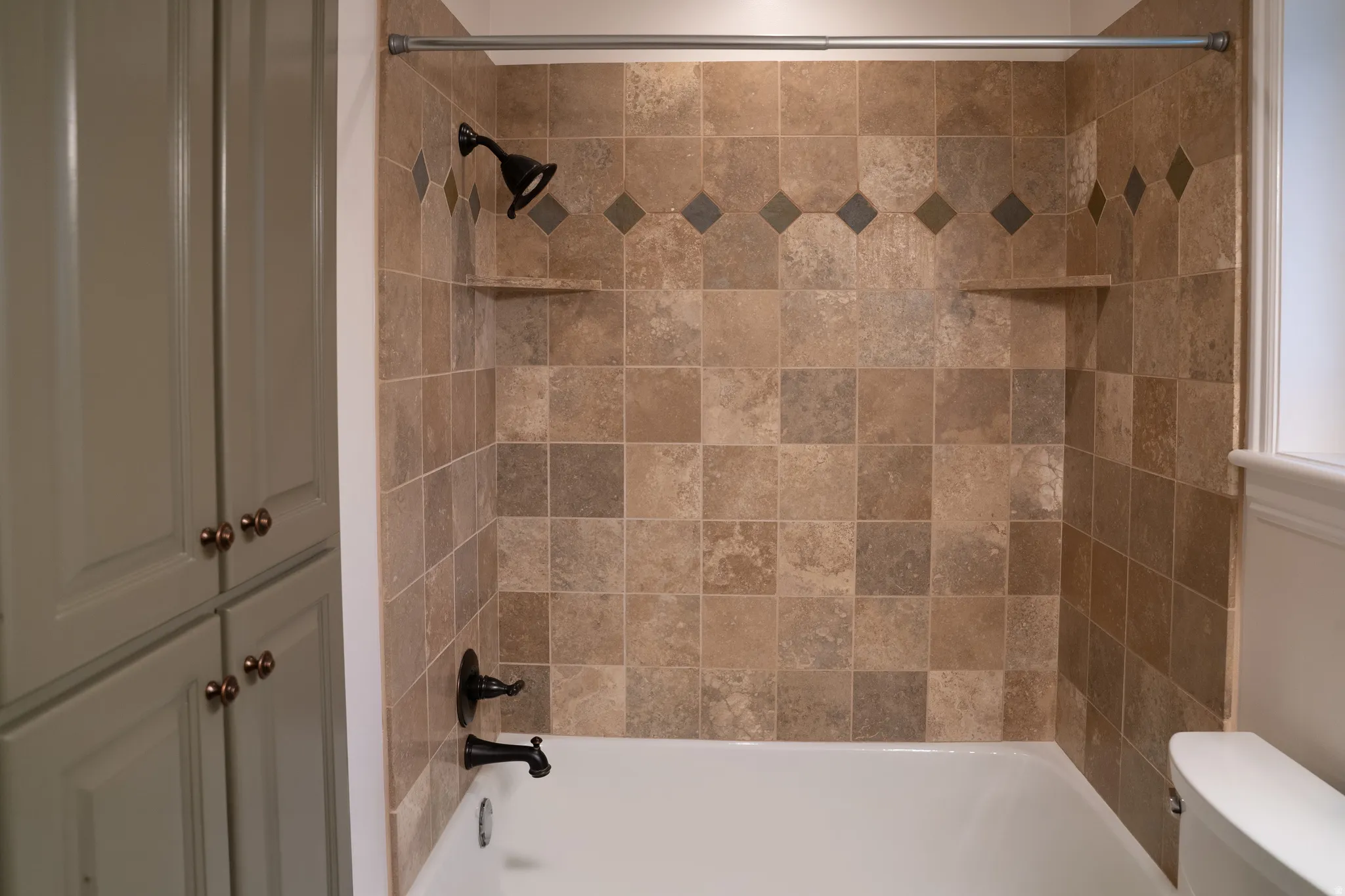 Bathtub shower with tiled surround featuring decorative accent course, built-in shelving, and dark bronze fixtures
