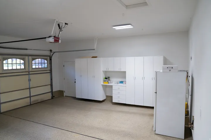 Spacious garage featuring a durable epoxy floor, comprehensive built-in white cabinetry, a dedicated workbench area, and an interior access door