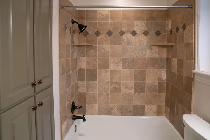 Bathtub shower with tiled surround featuring decorative accent course, built-in shelving, and dark bronze fixtures