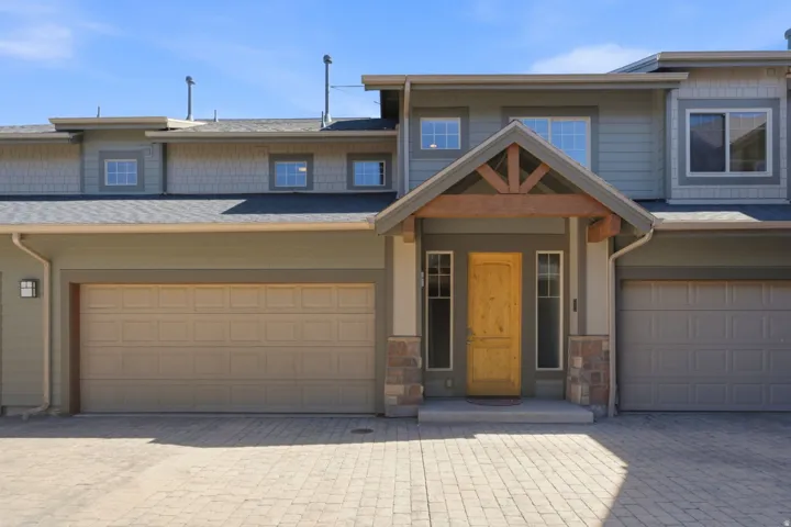 Exterior featuring a natural wood-finish door, stone accents, shingle siding, a covered entry with exposed wood beams, and a paver driveway