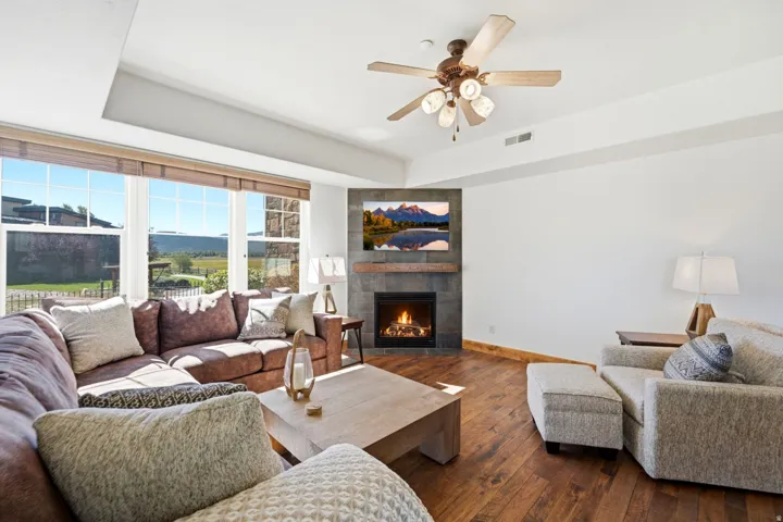Living room featuring an integrated fireplace with stone-finish surround, wood-finish flooring, a ceiling fan, and large windows