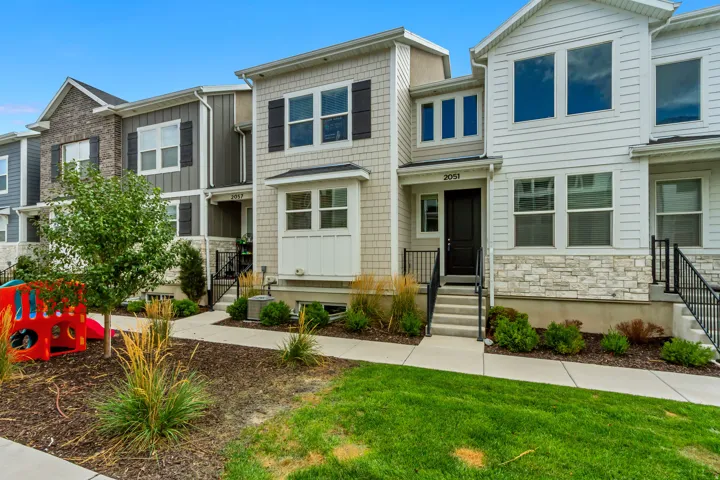 Multi-level townhouse exterior featuring light-colored siding, dark window shutters, a stone accent facade, a front walkway, and maintained landscaping