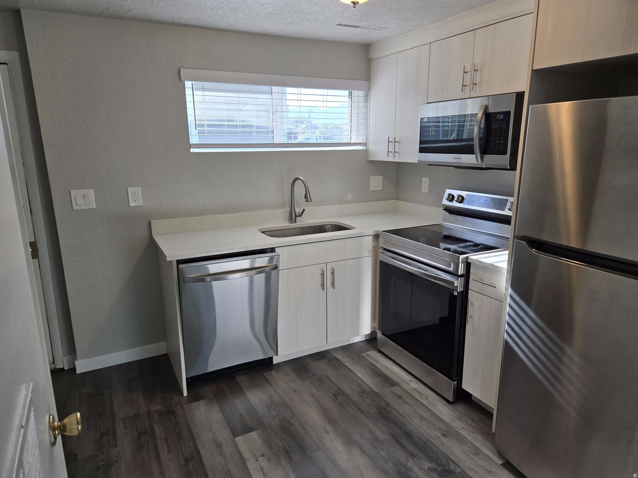 Modern kitchen featuring stainless steel appliances, light wood-finish cabinetry, white countertops, and wood-finish flooring