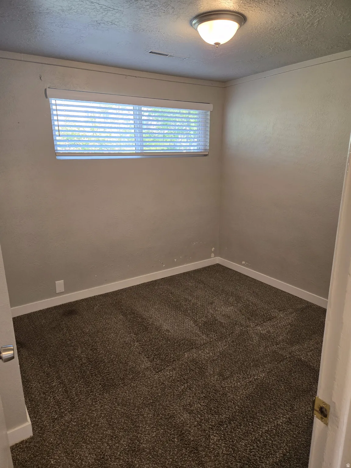 Room featuring neutral-toned walls, textured carpet flooring, and a single window with horizontal blinds