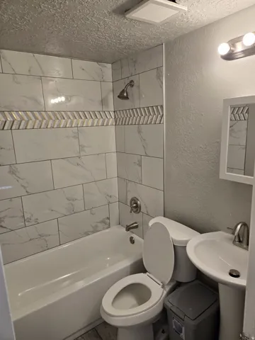 Bathtub with tile surround, decorative accent tile band, pedestal sink, and wall-mounted vanity lighting