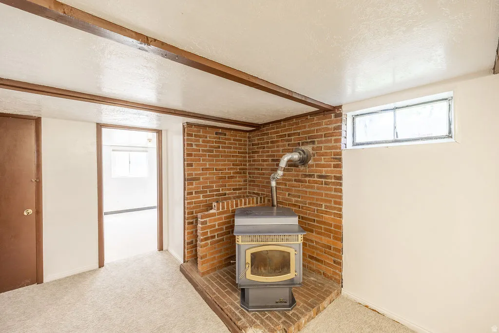 Brick wood-burning stove with metal flue, brown wood ceiling beams, neutral wall paint, and light-toned carpet flooring