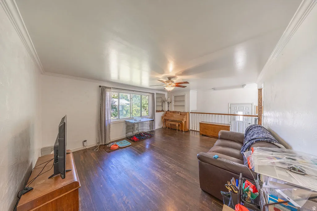 Hardwood-finish flooring, white painted walls, crown molding, ceiling fan, and large window