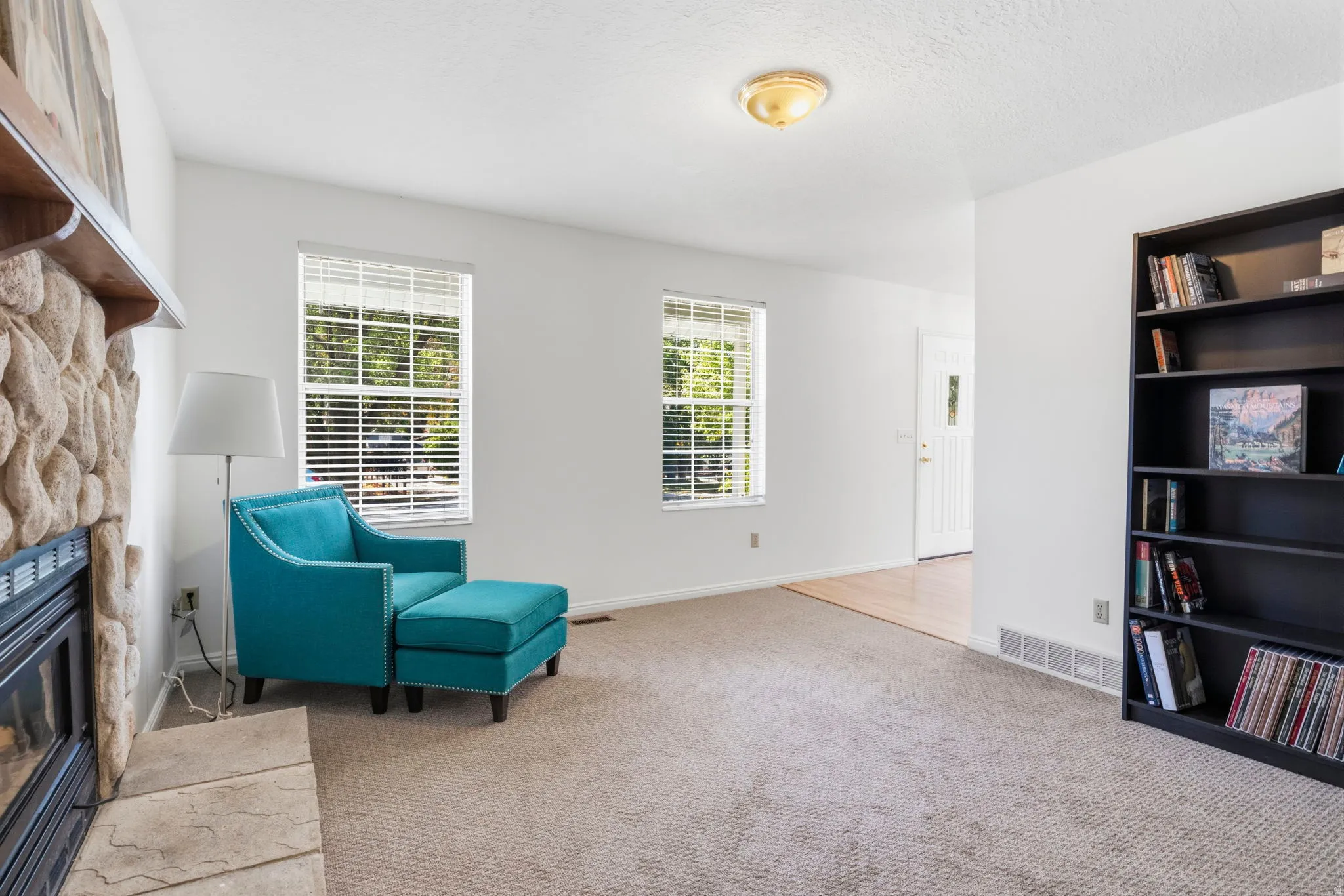 Carpeted living area featuring a stone-clad fireplace with mantel, multiple windows, a built-in bookshelf, and light wood-finish flooring