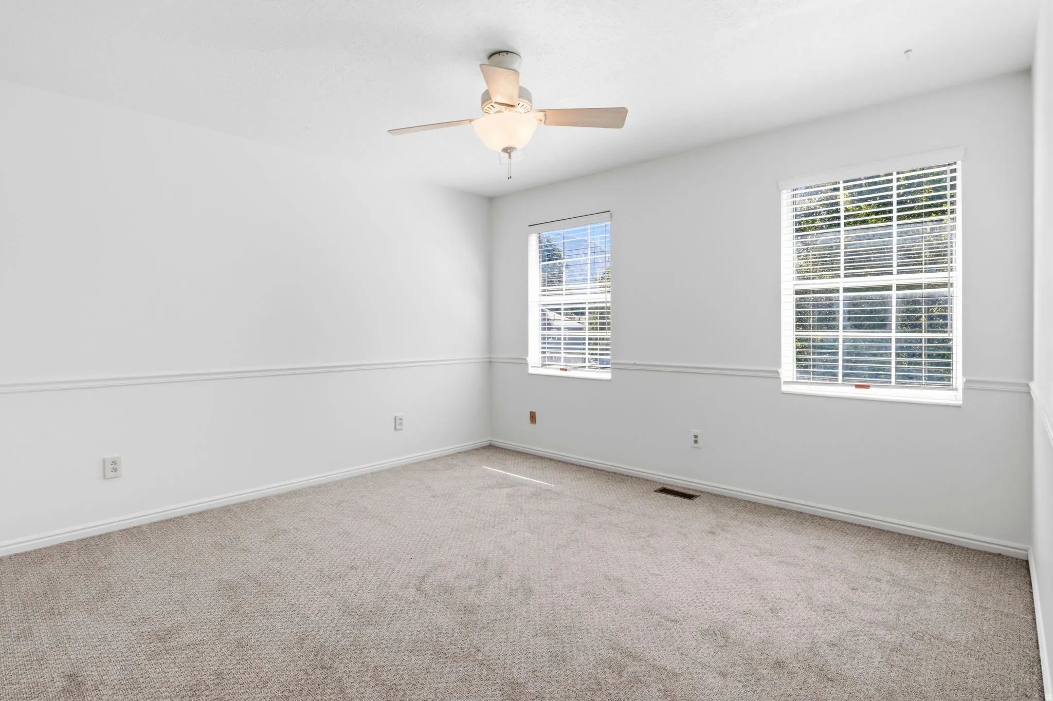 Plush carpet flooring, white painted walls, chair rail molding, two windows with blinds, and a ceiling fan with integrated lighting