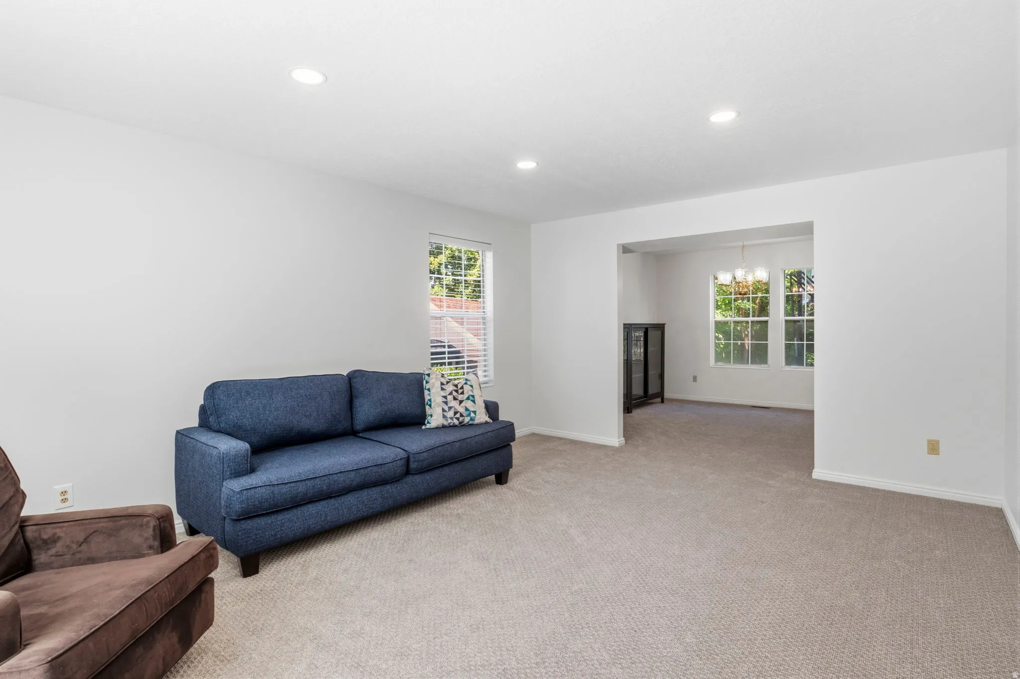 Recessed lighting illuminates the carpeted living area with white walls and a wide archway to an additional room