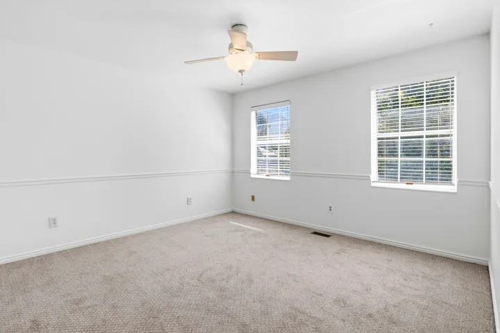 Plush carpet flooring, white painted walls, chair rail molding, two windows with blinds, and a ceiling fan with integrated lighting
